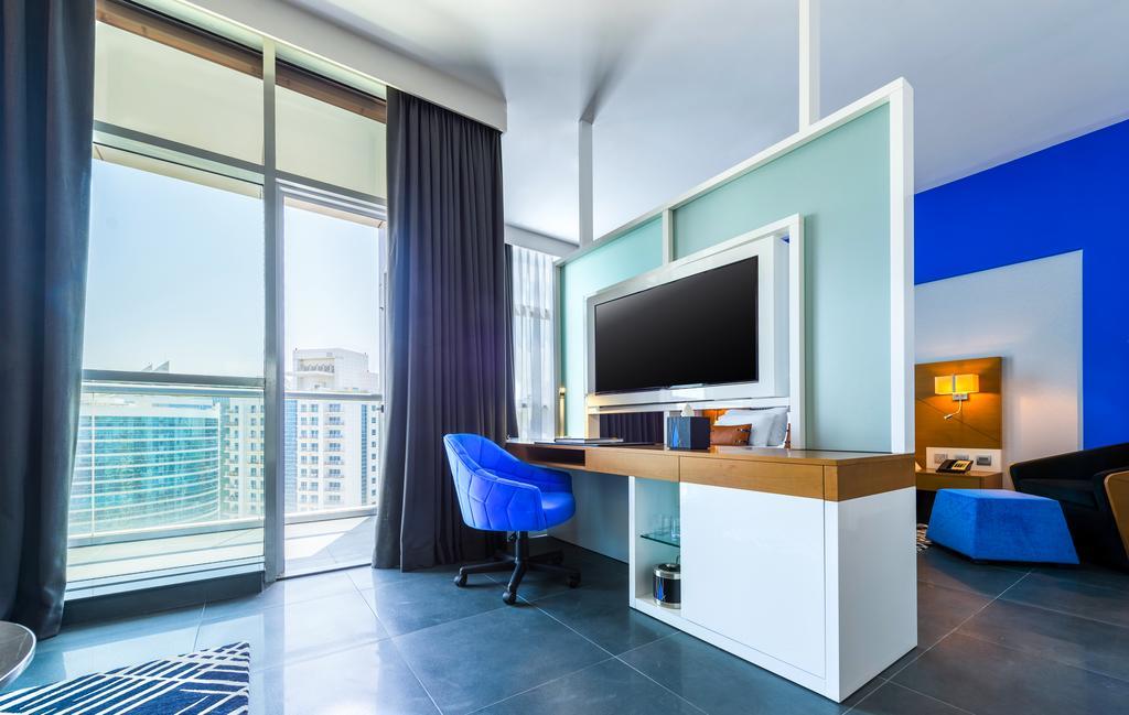 Tryp By Wyndham Dubai Hotel Ngoại thất bức ảnh The photo shows a modern room with large windows allowing natural light to enter. There's a stylish desk area featuring a flat-screen TV mounted on the wall, along with a light wooden desk and a blue chair. The wall behind the TV is painted in a ligh